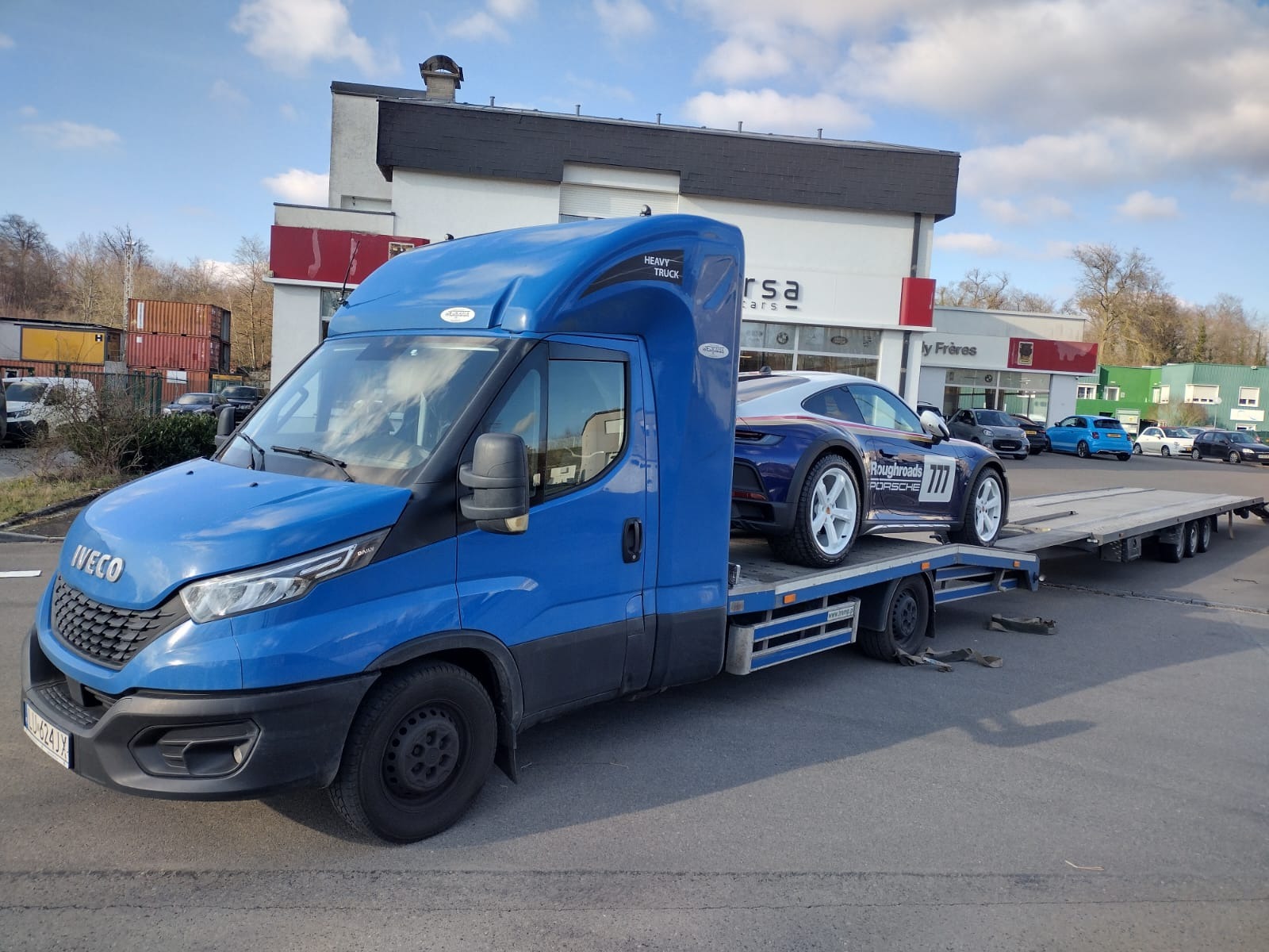 Niebieska laweta Iveco z załadowanym sportowym Porsche z białymi felgami na platformie, zaparkowana na tle budynku z logo firmy motoryzacyjnej.