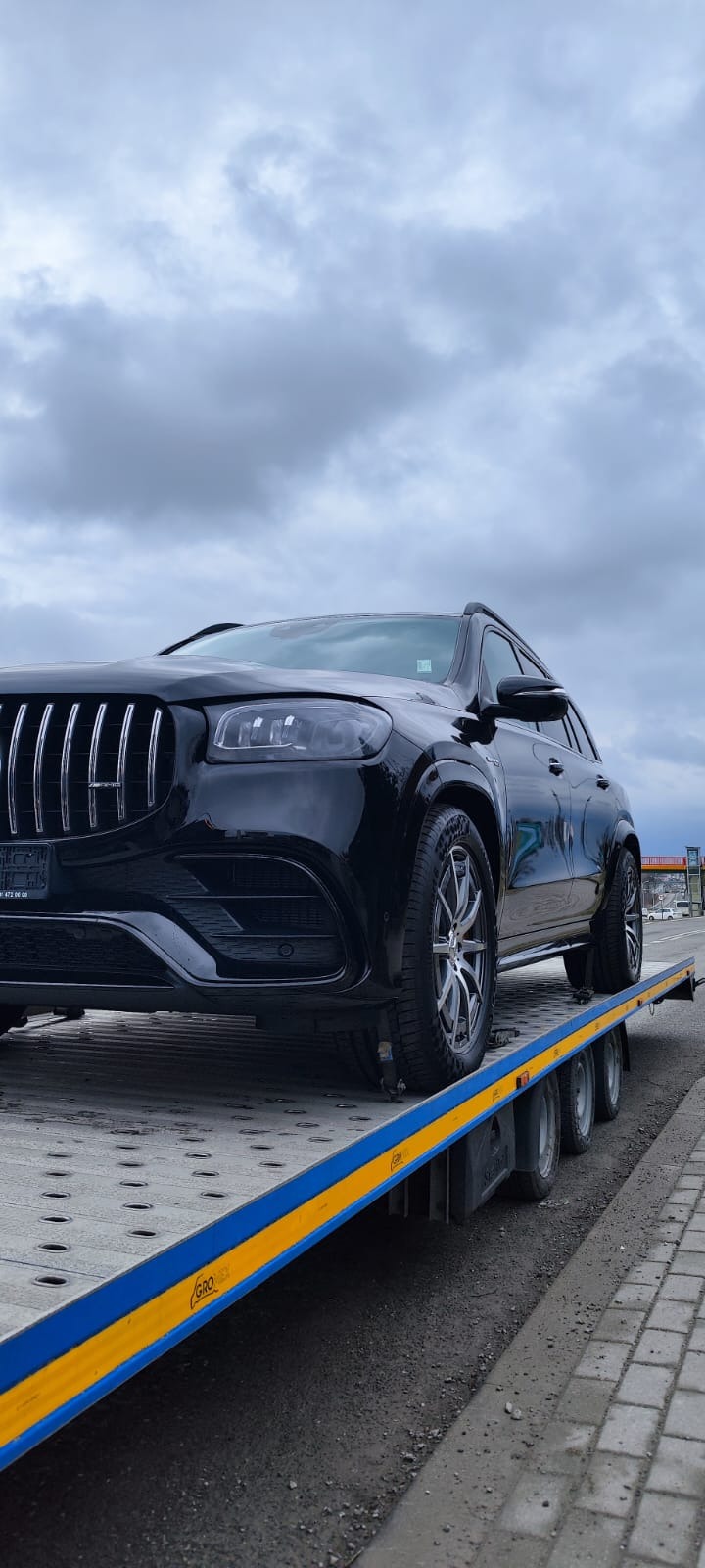 Czarny SUV Mercedes-Benz na platformie lawety z niebiesko-żółtymi krawędziami, pod pochmurnym niebem.