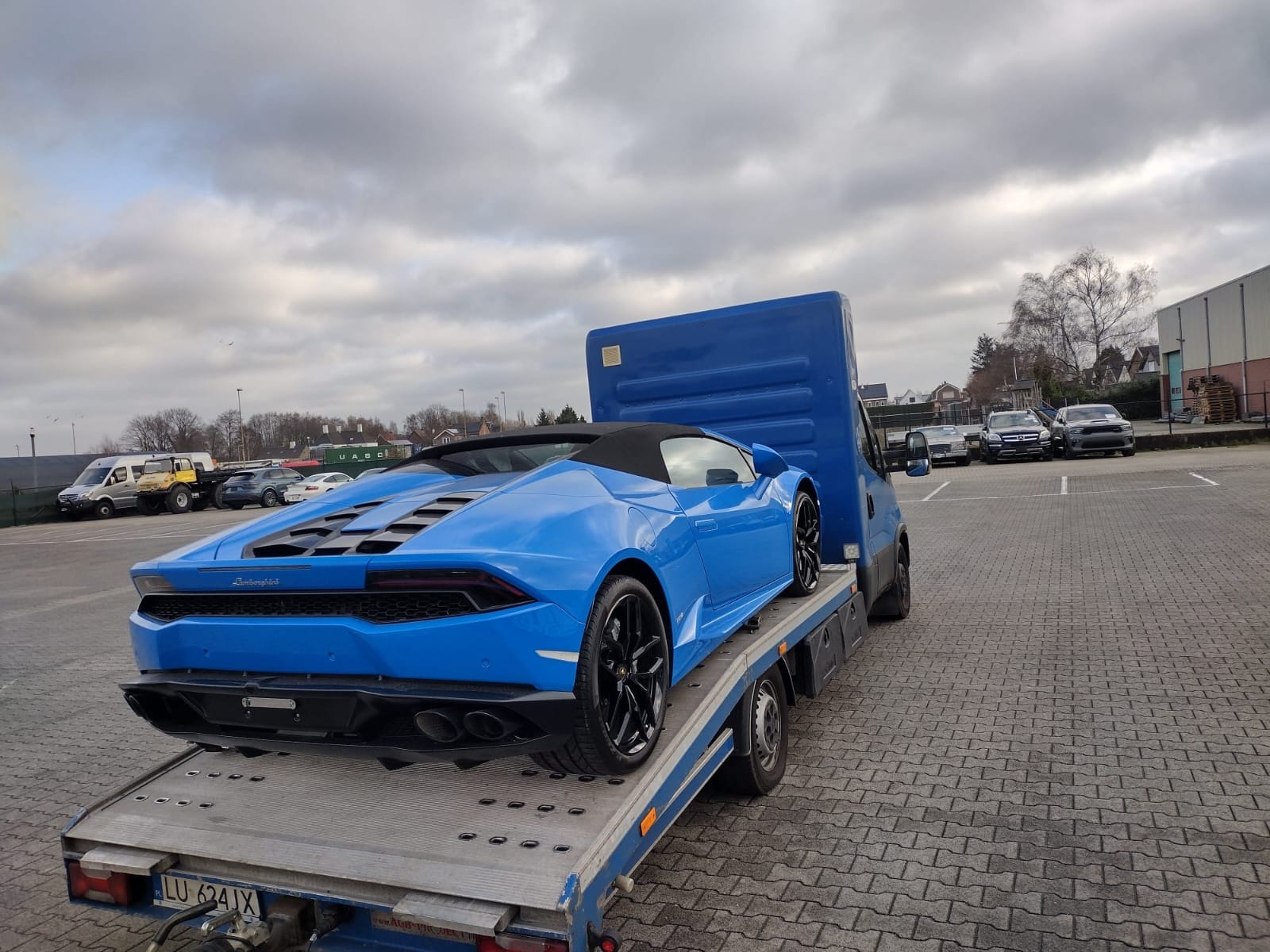 Błękitny Lamborghini Huracan Spyder na niebieskiej lawecie z widoczną tablicą rejestracyjną, na tle parkingu z innymi samochodami i pochmurnym niebem.
