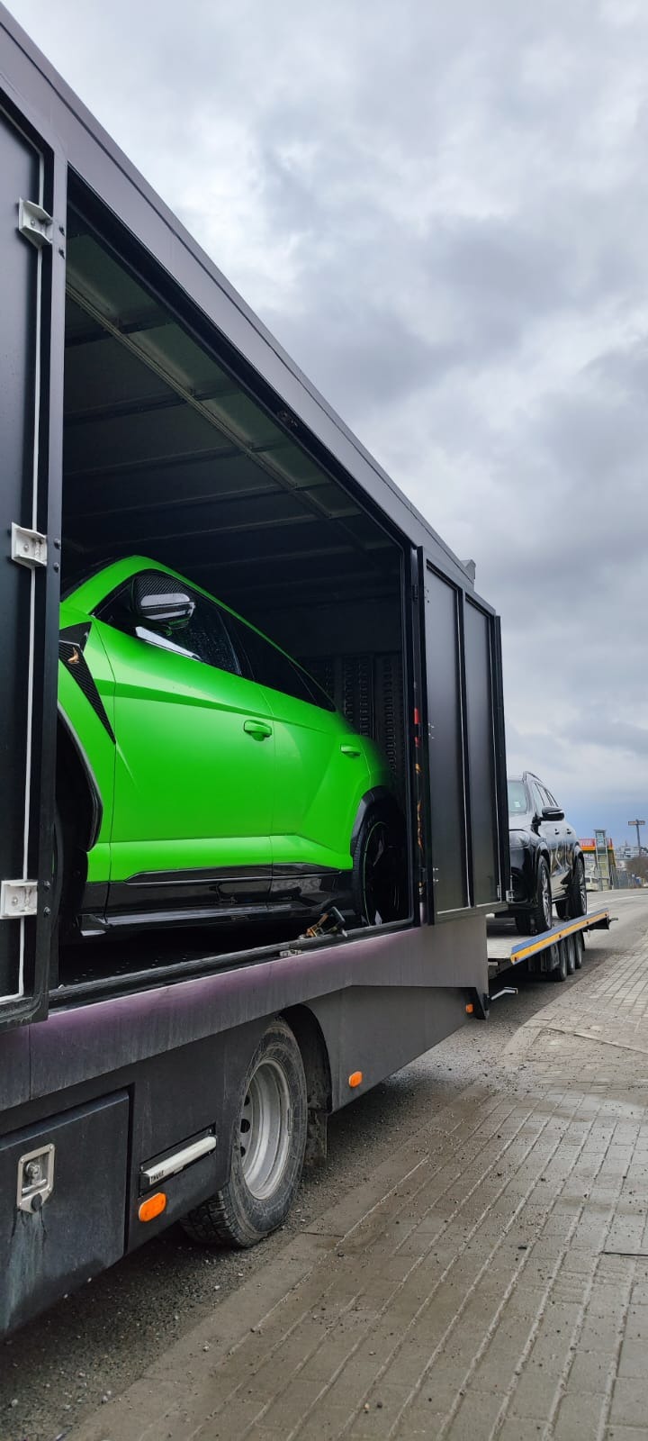 Czarna laweta z otwartą przestrzenią ładunkową, w której znajduje się jaskrawozielony samochód sportowy oraz czarny SUV na platformie z tyłu, na tle pochmurnego nieba.