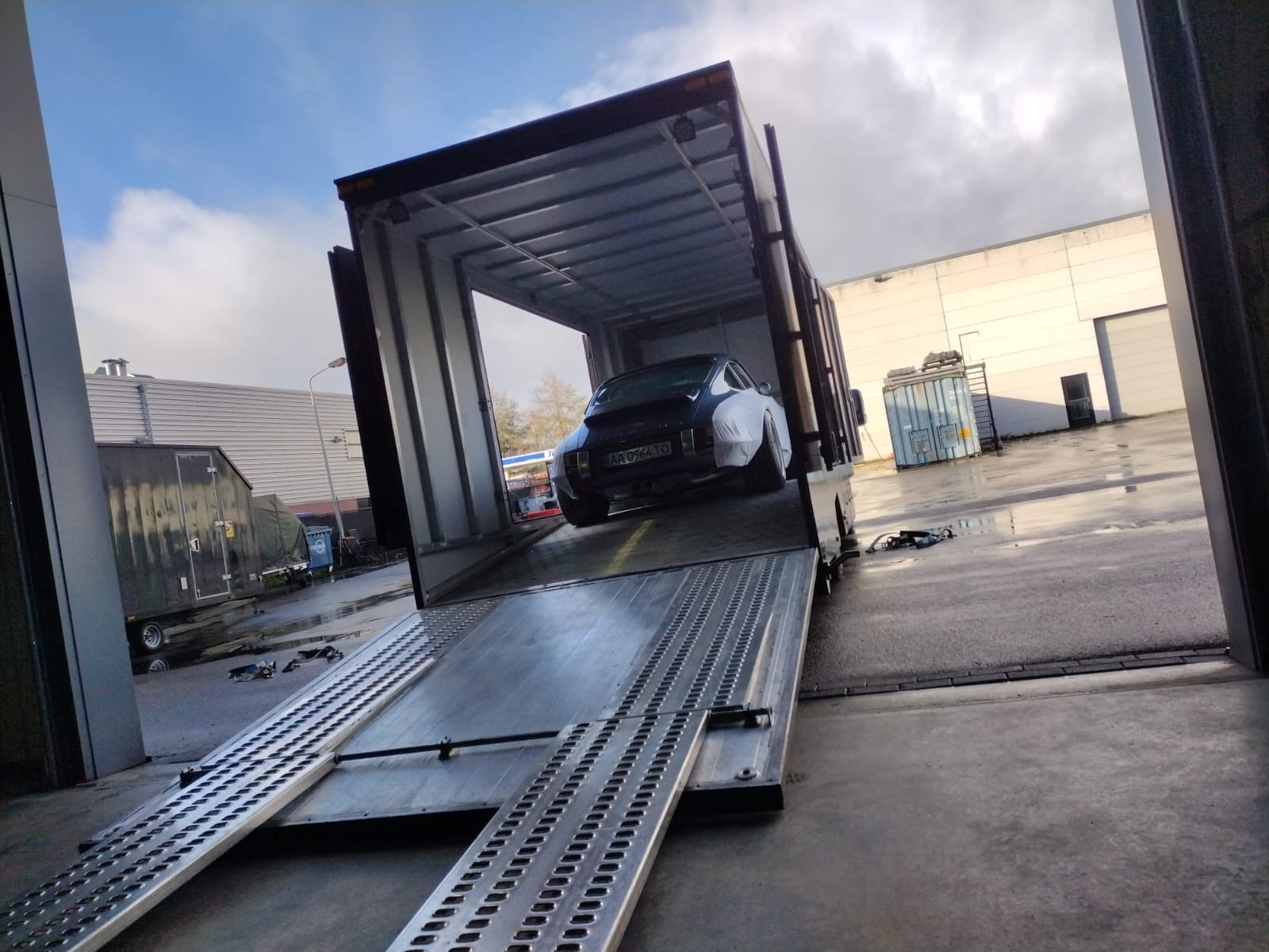 Czarny samochód sportowy, częściowo owinięty folią ochronną, wjeżdża na platformę lawety z metalowymi najazdami, na mokrym placu przemysłowym w pochmurny dzień.