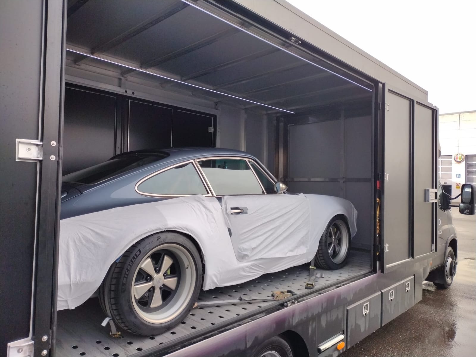 Szary samochód sportowy, częściowo przykryty białą folią ochronną, wewnątrz zamkniętej, oświetlonej lawety z metalową podłogą kratową i czarnymi ścianami, widoczny fragment kabiny lawety.