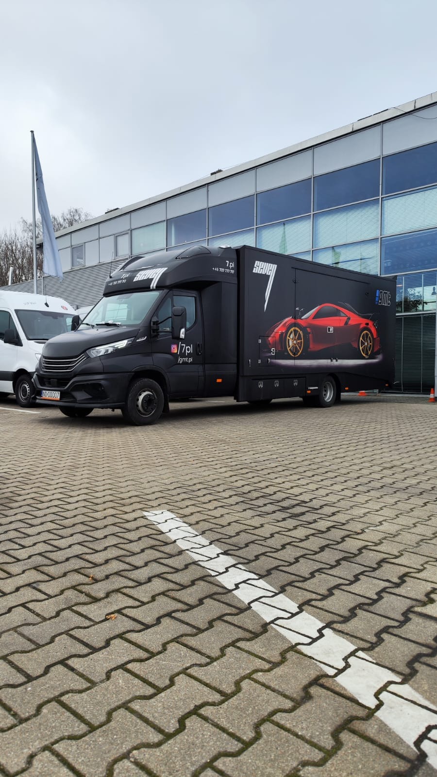 Czarna laweta z grafiką czerwonego sportowego samochodu na boku, zaparkowana na brukowanym parkingu przed nowoczesnym budynkiem biurowym.