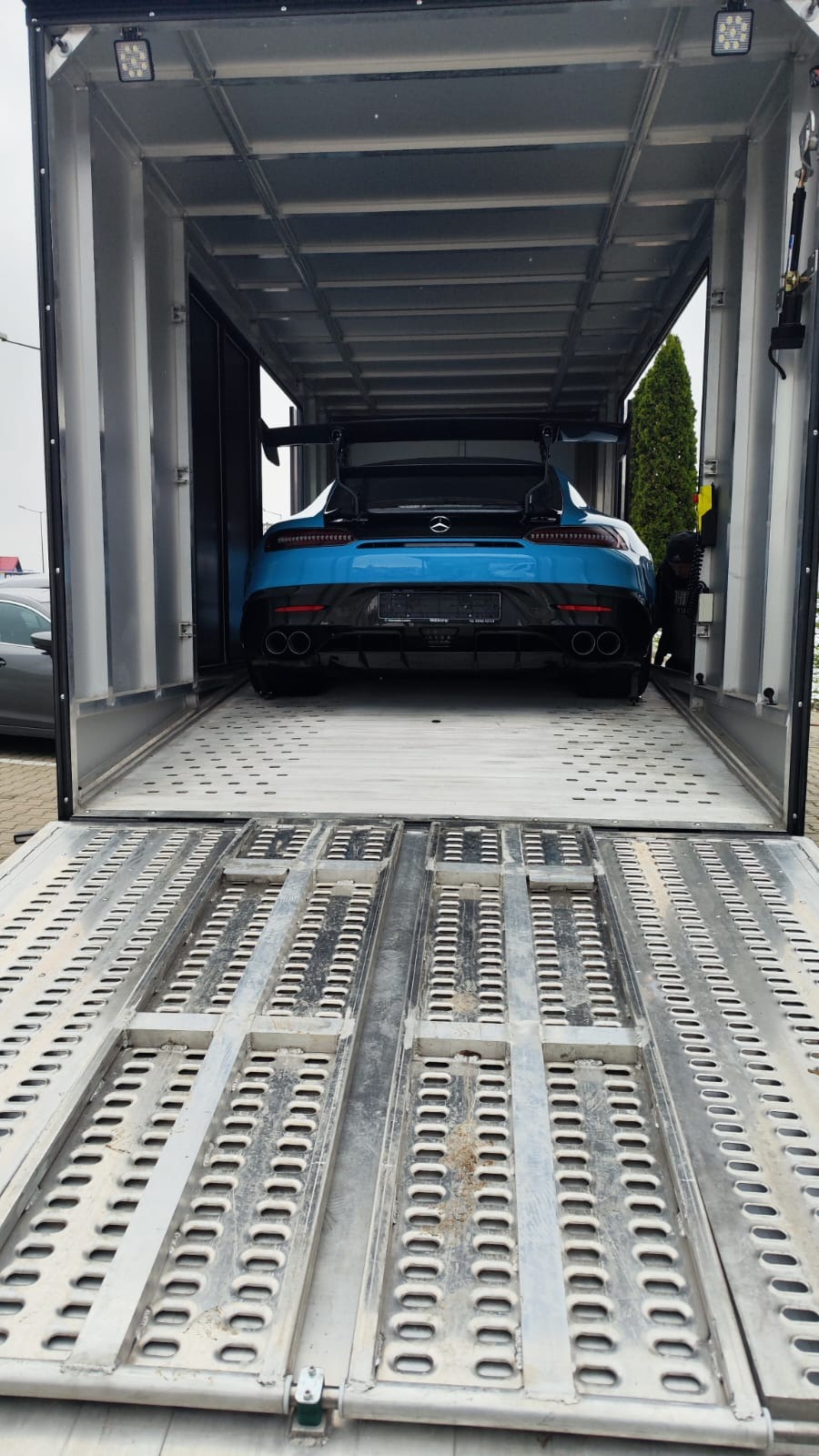 Błękitny sportowy samochód Mercedes-AMG GT z dużym spojlerem, umieszczony wewnątrz metalowej lawety z otwartą rampą, widok z tyłu.