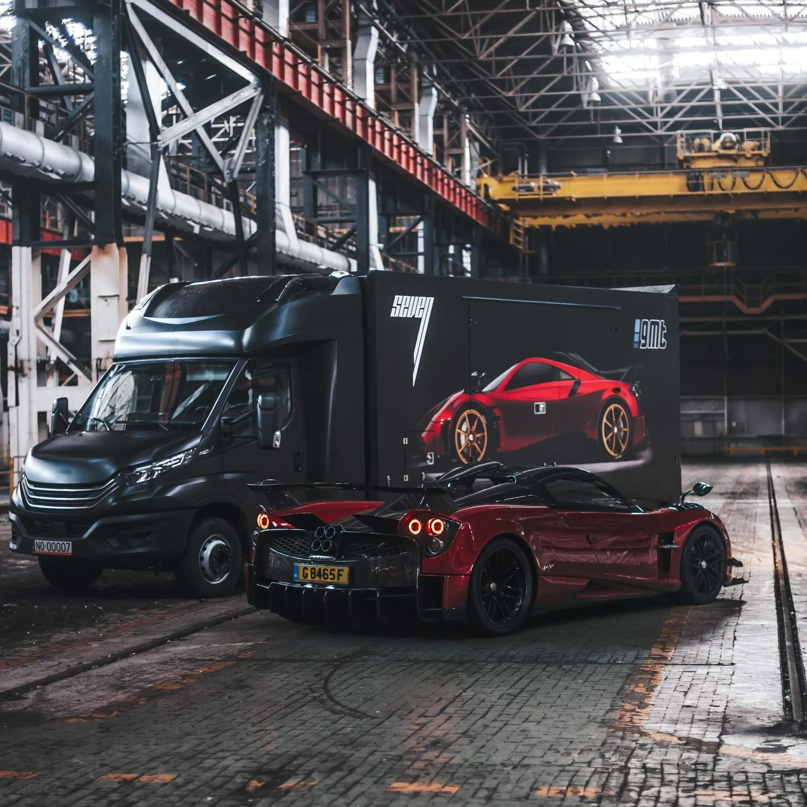 Czarny bus dostawczy z grafiką czerwonego sportowego samochodu obok rzeczywistego czerwonego samochodu sportowego w hali przemysłowej.