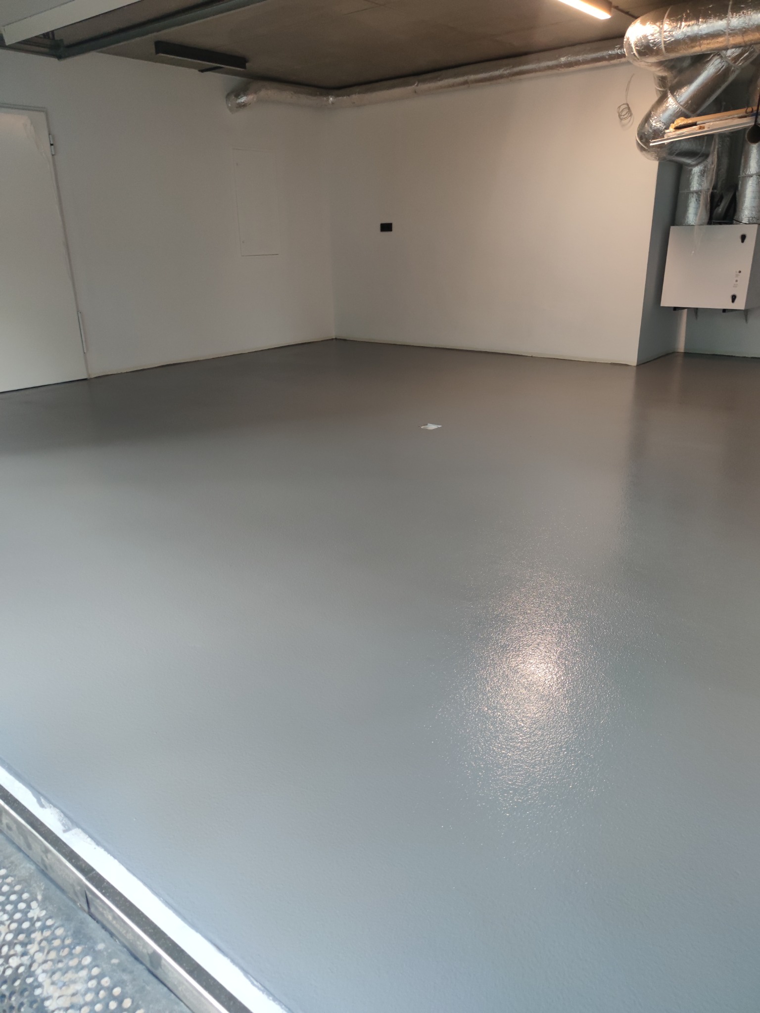 Szara, gładka posadzka żywiczna w garażu z widocznymi rurami wentylacyjnymi pod sufitem i białą ścianą z drzwiami po lewej stronie.