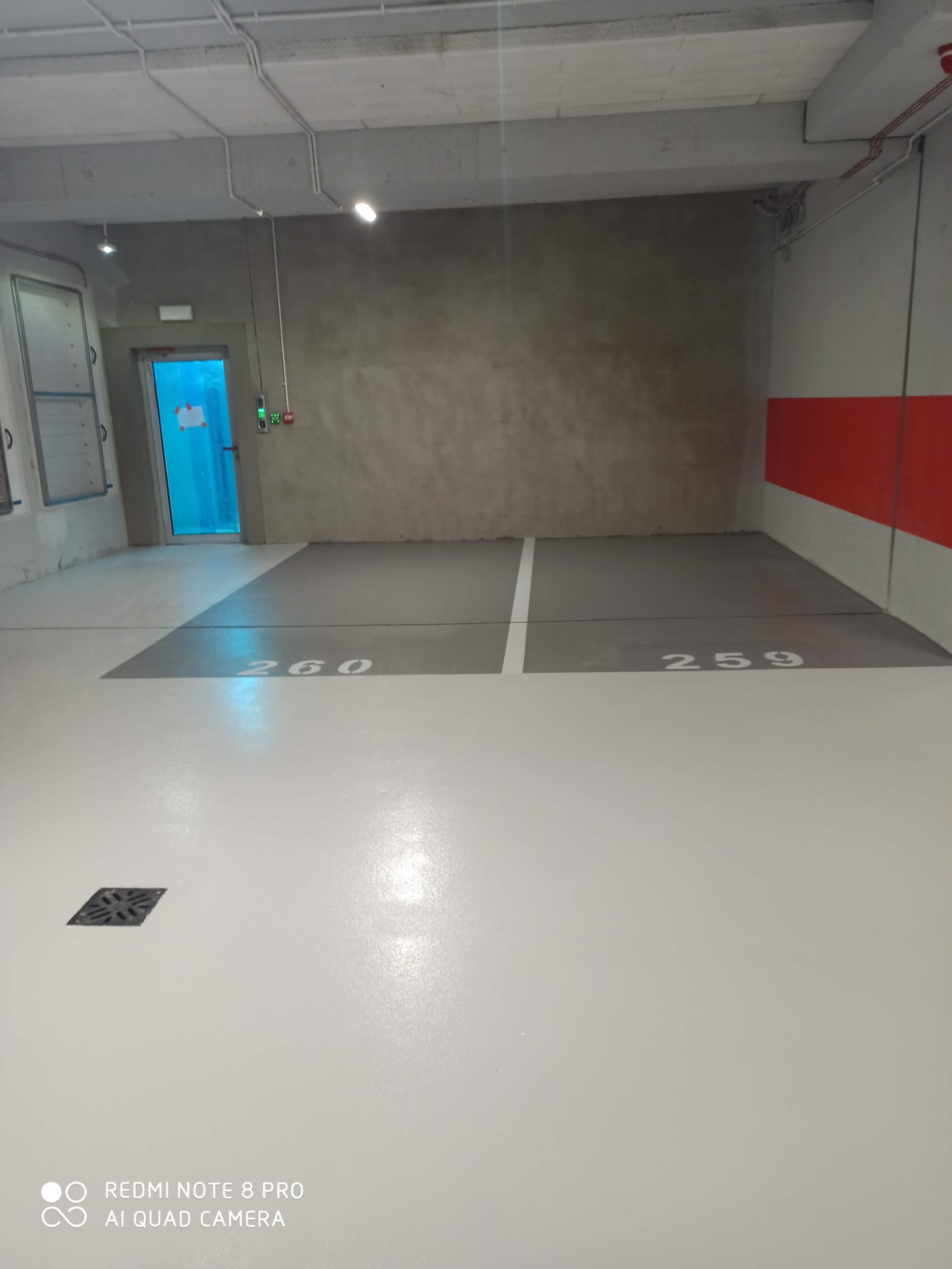 Biała posadzka w garażu podziemnym z szarymi numerami miejsc parkingowych 260 i 259, widoczny odpływ liniowy i drzwi ewakuacyjne.