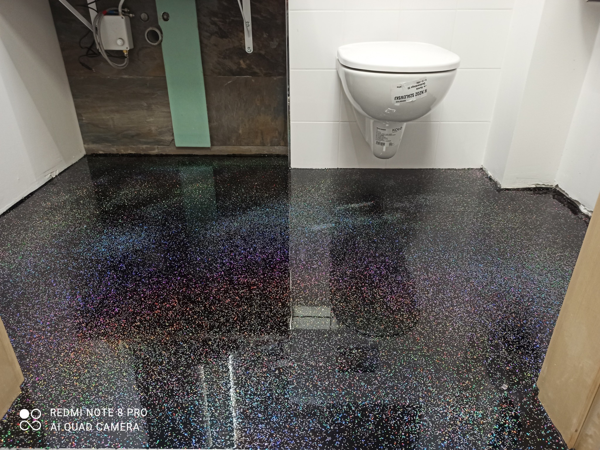 Błyszcząca, ciemna podłoga w łazience z brokatowym efektem, widoczna muszla klozetowa i fragment instalacji wodnej.