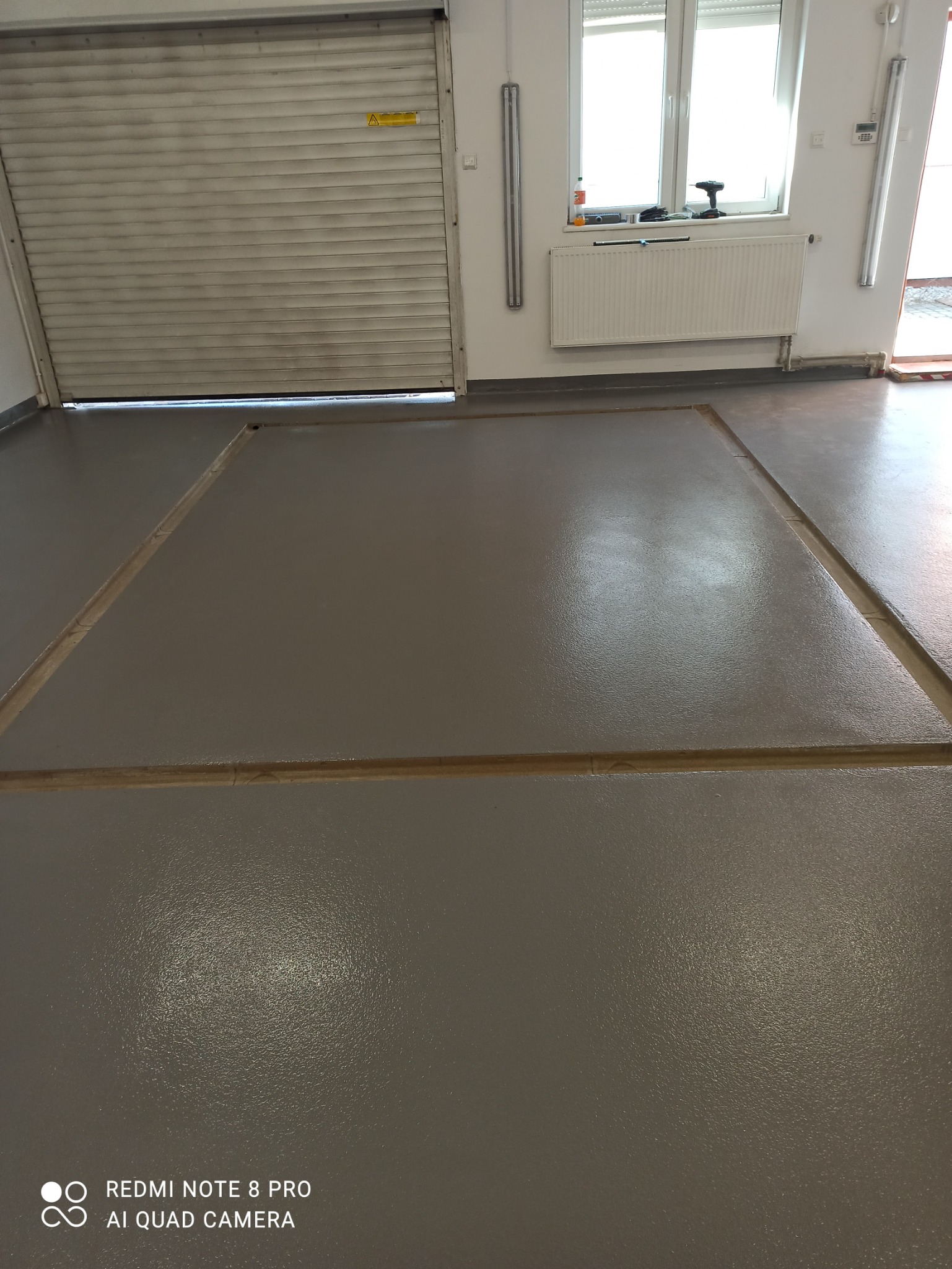 Szara, gładka posadzka w garażu lub hali, podzielona listwami dylatacyjnymi, z widoczną bramą garażową i oknem w tle.