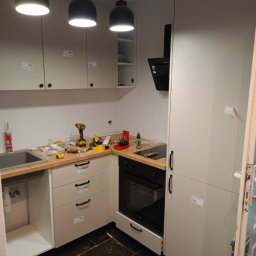 Hubert Olczak - Montaż szarych szafek kuchennych z drewnianym blatem, widoczna wiertarka, narzędzia i okap, czarne lampy wiszące, ciemna podłoga. Częściowo zamontowana kuchnia.