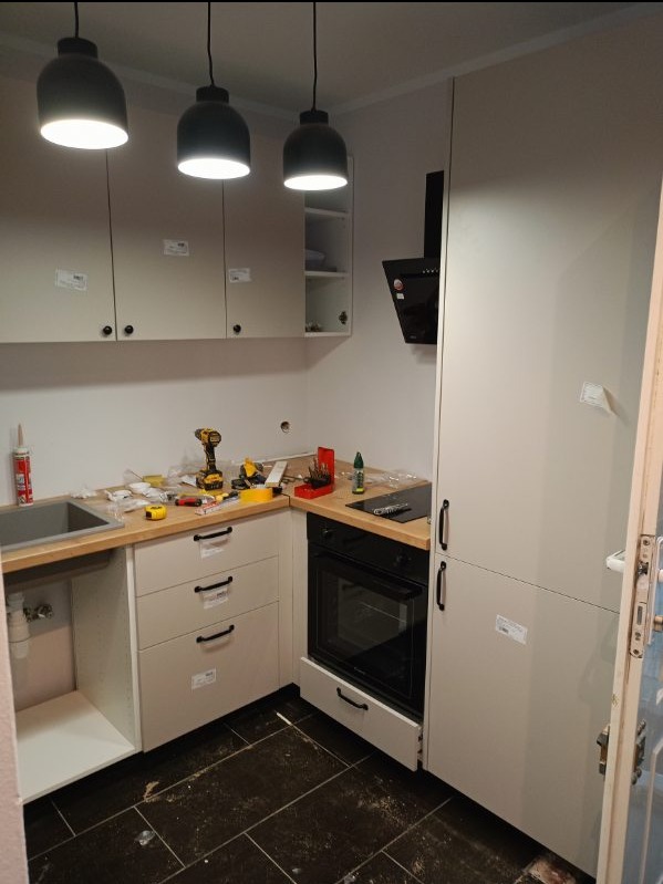 Montaż szarych szafek kuchennych z drewnianym blatem, widoczna wiertarka, narzędzia i okap, czarne lampy wiszące, ciemna podłoga. Częściowo zamontowana kuchnia.