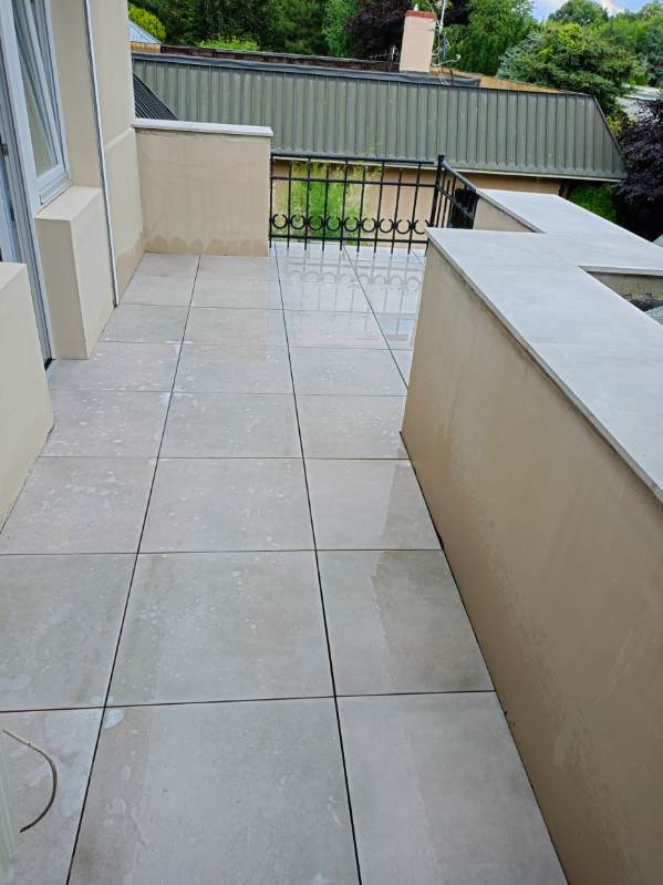 Wyłożony jasnymi, kwadratowymi płytkami balkon z beżowymi ścianami i czarną, metalową balustradą, widok na zielone drzewa i fragment dachu budynku w tle.