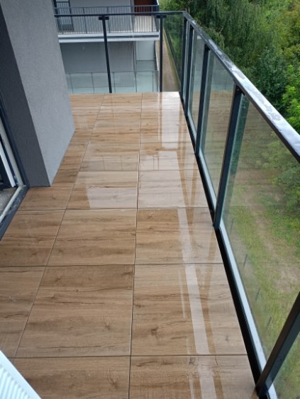Błyszczące płytki imitujące drewno na balkonie z szklaną balustradą i widokiem na zieleń.