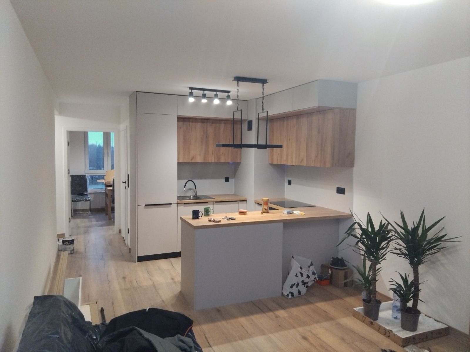 Jasne wnętrze z aneksem kuchennym w trakcie prac wykończeniowych, widoczne niedoskonałości na podłodze, rośliny doniczkowe w rogu pomieszczenia, nowoczesne oświetlenie nad wyspą kuchenną.
