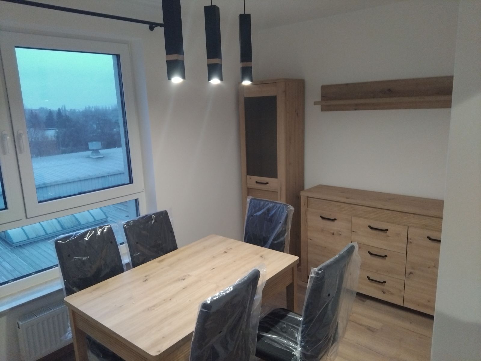 Jasne drewniane meble w nowym mieszkaniu: stół z krzesłami w folii ochronnej, komoda i witryna, oświetlenie punktowe.