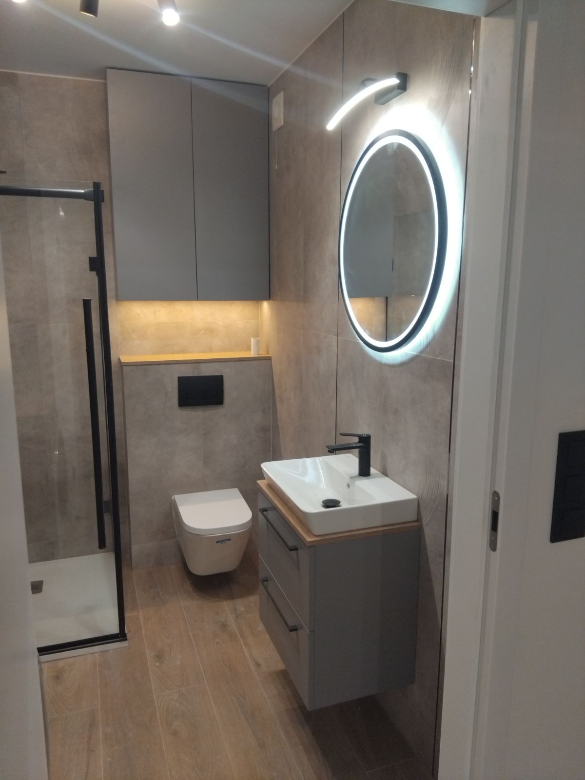Nowoczesna łazienka z kabiną prysznicową typu walk-in, podwieszaną toaletą, szarą szafką pod umywalką z drewnianym blatem i okrągłym lustrem z podświetleniem LED.