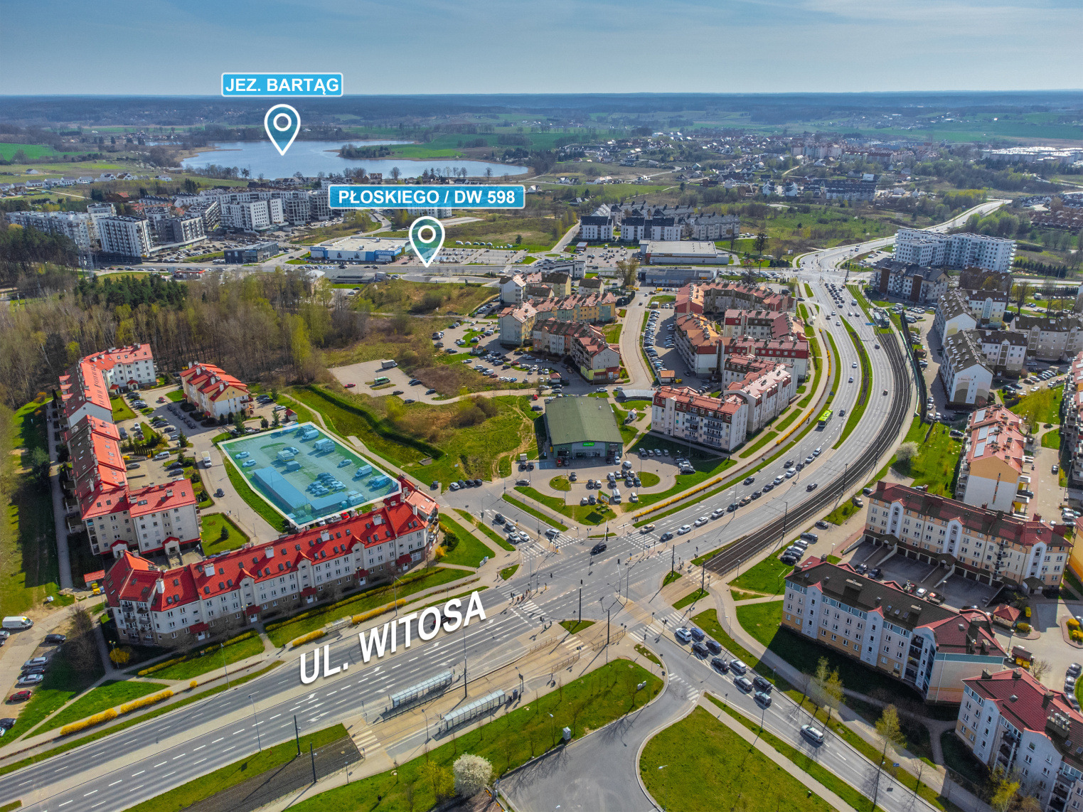 Zdjęcie z drona przedstawiające widok na osiedle mieszkaniowe z czerwonymi dachami, ulicę Witosa, jezioro Bartąg i drogę Płoskiego/DW 598 w słoneczny dzień.