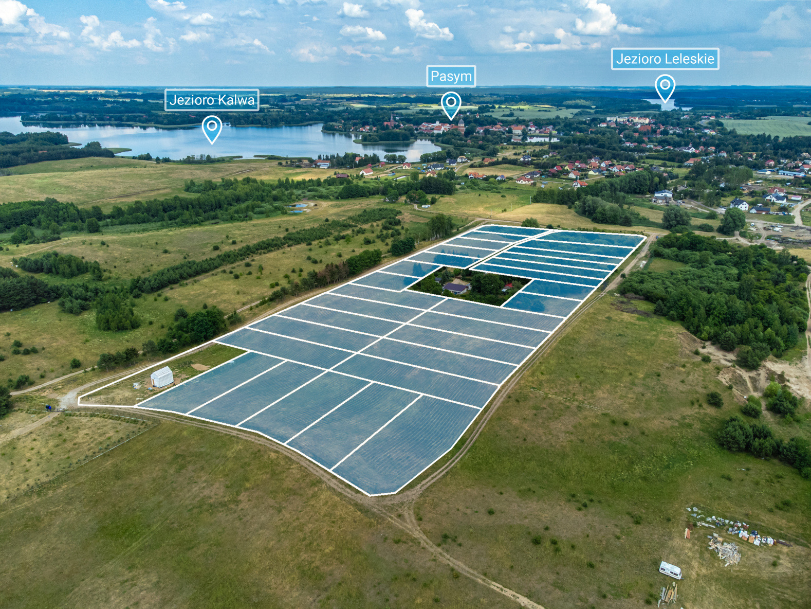 Panoramiczny widok z lotu ptaka na farmę fotowoltaiczną zlokalizowaną w pobliżu jezior i zabudowań miejskich, otoczoną zielonymi polami i lasami.