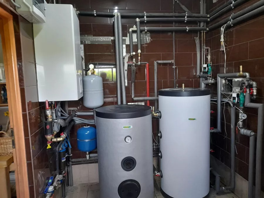 Kotłownia z dwoma zbiornikami akumulacyjnymi, piecem gazowym i rozbudowaną instalacją hydrauliczną z rur miedzianych i zaworów, widok z bliska na brązowe kafelki ścienne.