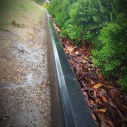 M DESING SPÓŁKA Z OGRANICZONĄ ODPOWIEDZIALNOŚCIĄ - Ukośne ujęcie czarnej obrzeża trawnika oddzielającego mokrą, błotnistą ziemię od kory drzewnej i gęstych, zielonych krzewów.