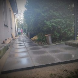 M DESING SPÓŁKA Z OGRANICZONĄ ODPOWIEDZIALNOŚCIĄ - Ułożona ścieżka z szarych, kwadratowych płyt betonowych prowadząca wzdłuż domu w kierunku zieleni, widoczne nierówności terenu i niedokończone fugowanie, osoba w oddali.