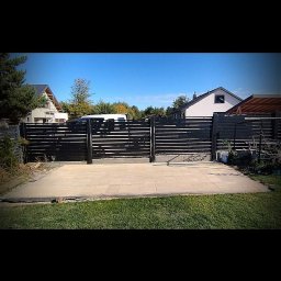 M DESING SPÓŁKA Z OGRANICZONĄ ODPOWIEDZIALNOŚCIĄ - Nowoczesne, poziome ogrodzenie panelowe w kolorze antracytowym, z widoczną kostką brukową na podjeździe, domy w tle.