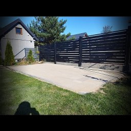 M DESING SPÓŁKA Z OGRANICZONĄ ODPOWIEDZIALNOŚCIĄ - Nowoczesne, poziome ogrodzenie z czarnych paneli, betonowy podjazd, trawnik i budynek w tle