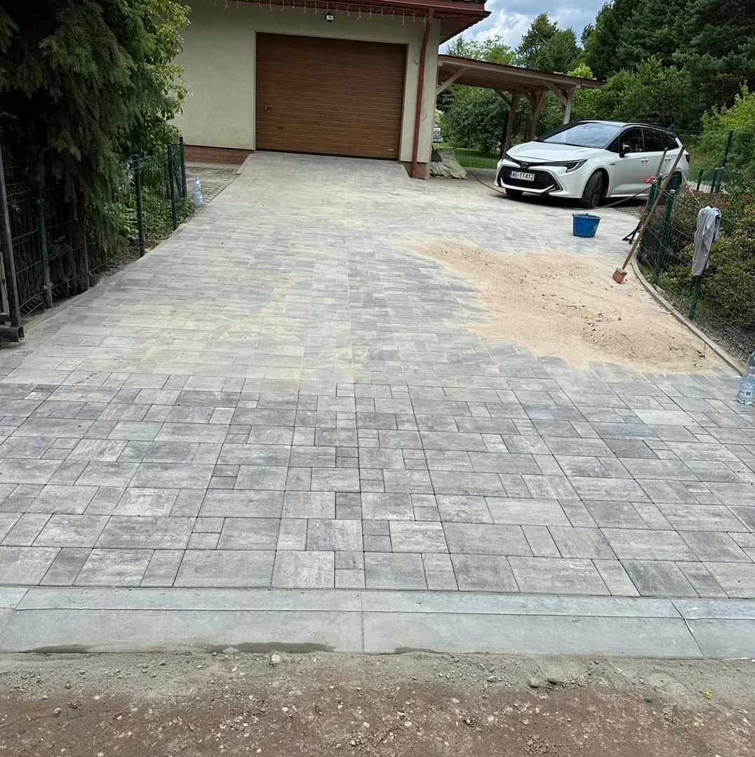 Ułożona szara kostka brukowa na podjeździe do garażu i domu, z widocznym fragmentem podsypki piaskowej, niebieskim wiadrem i białym samochodem w tle.