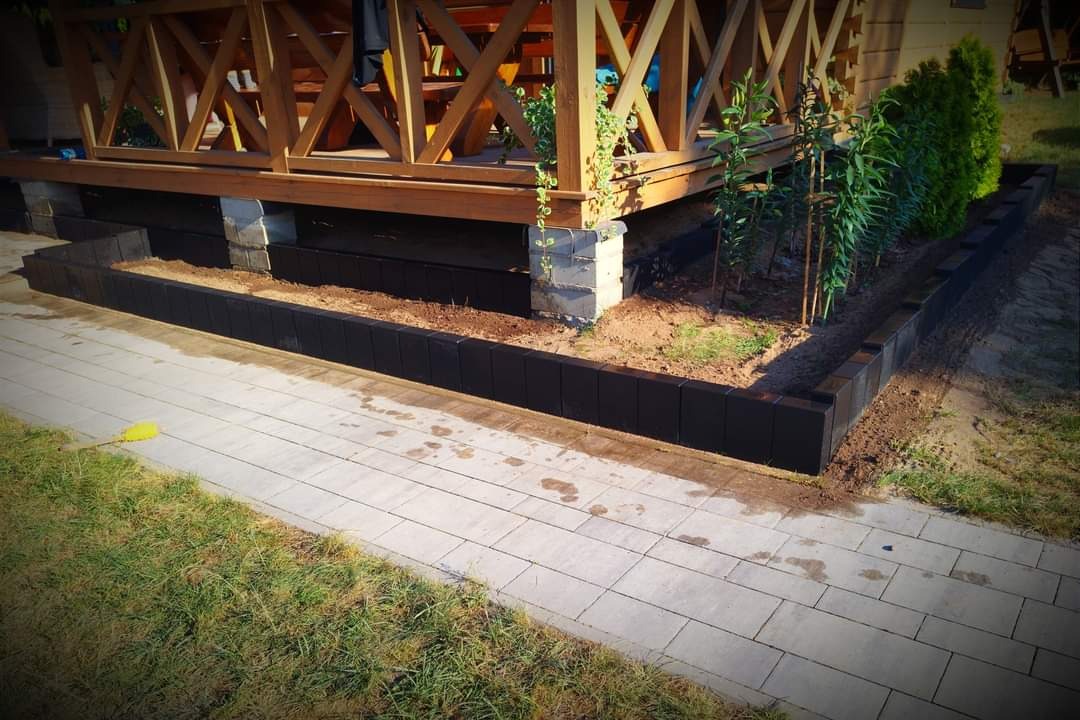 Obrzeże rabaty z czarnych kostek betonowych, oddzielające drewnianą konstrukcję tarasu od ścieżki z jasnej kostki brukowej, z młodymi roślinami wewnątrz.