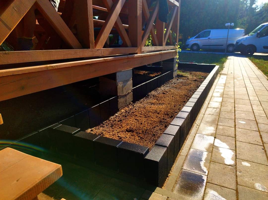 Wykończona rabata kwiatowa obłożona czarną kostką brukową, widok z boku, w tle drewniana konstrukcja i zaparkowane samochody dostawcze.