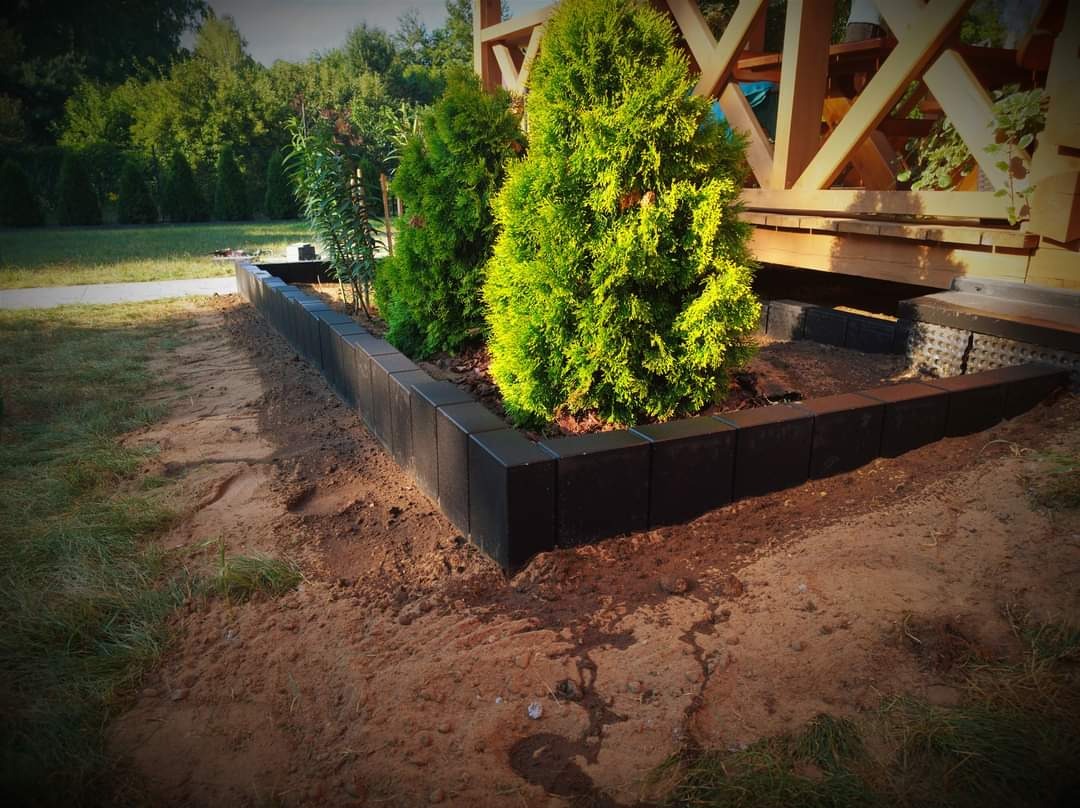 Krawężnik z ciemnej kostki obramowujący rabatę z tujami przy drewnianej konstrukcji altany, ziemia wokół rabaty.