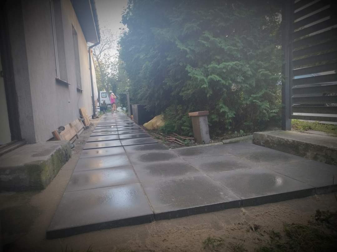 Ułożona ścieżka z szarych, kwadratowych płyt betonowych prowadząca wzdłuż domu w kierunku zieleni, widoczne nierówności terenu i niedokończone fugowanie, osoba w oddali.