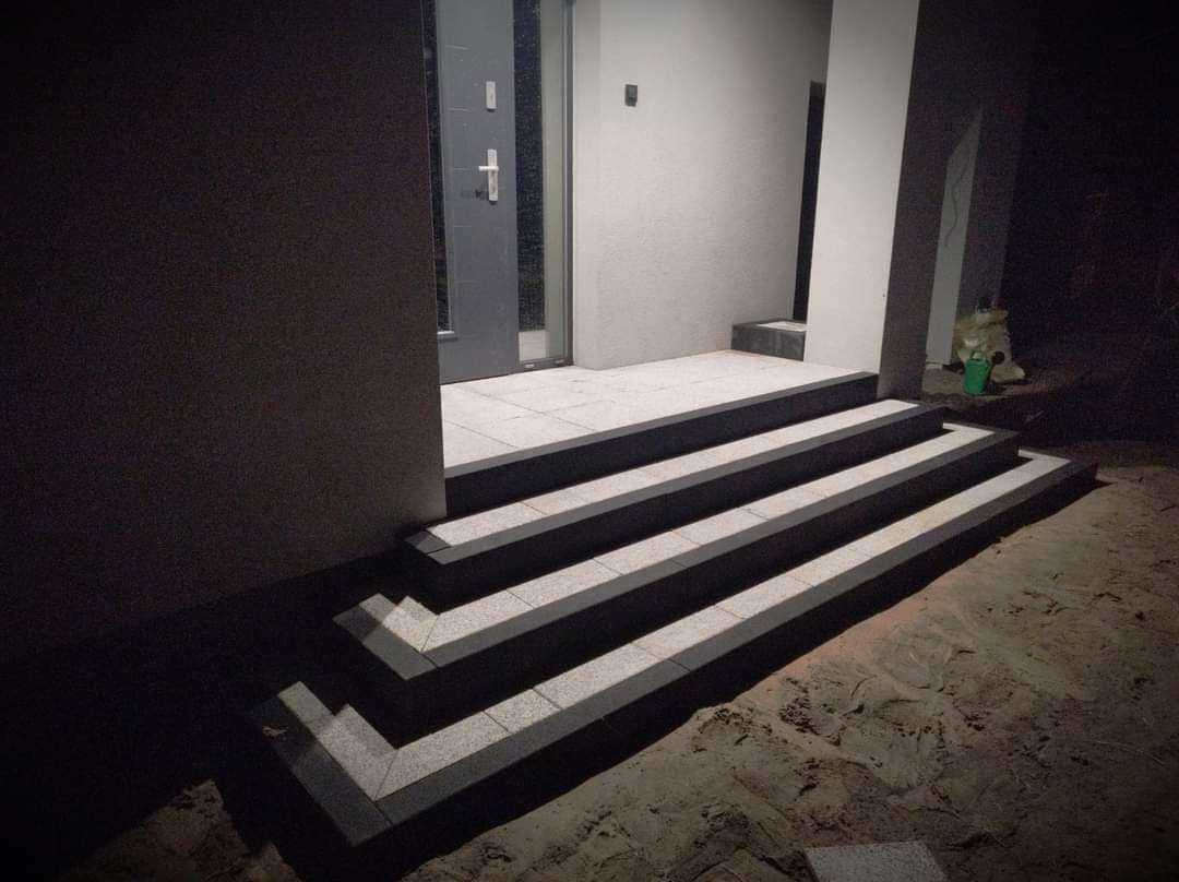 Nowoczesne schody wejściowe wyłożone jasną kostką brukową z ciemnymi obrzeżami, prowadzące do szarych drzwi wejściowych w budynku o jasnej elewacji, widoczne w słabym oświetleniu nocnym.