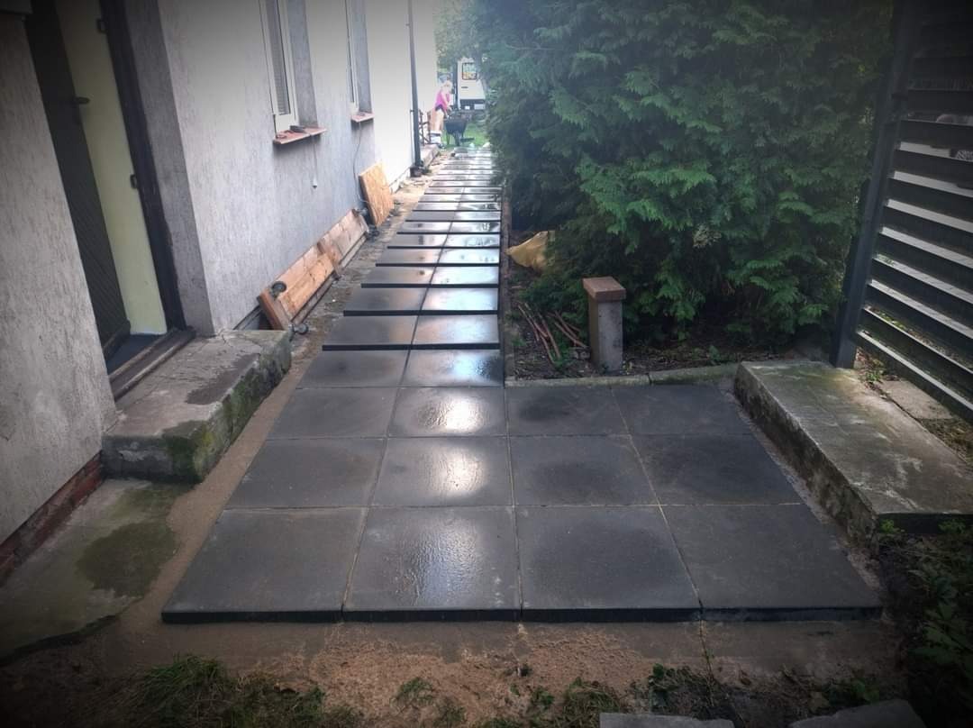 Ułożona ścieżka z ciemnych, kwadratowych płyt betonowych prowadząca wzdłuż budynku, z widocznymi przerwami między płytami wypełnionymi piaskiem, oraz przygotowanym podłożem pod dalszą część ścieżki.