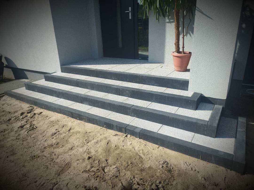 Schody wejściowe wyłożone jasnoszarą kostką brukową z ciemnoszarym obrzeżem, prowadzące do drzwi domu w Grodzisku Mazowieckim, obok donica z rośliną.