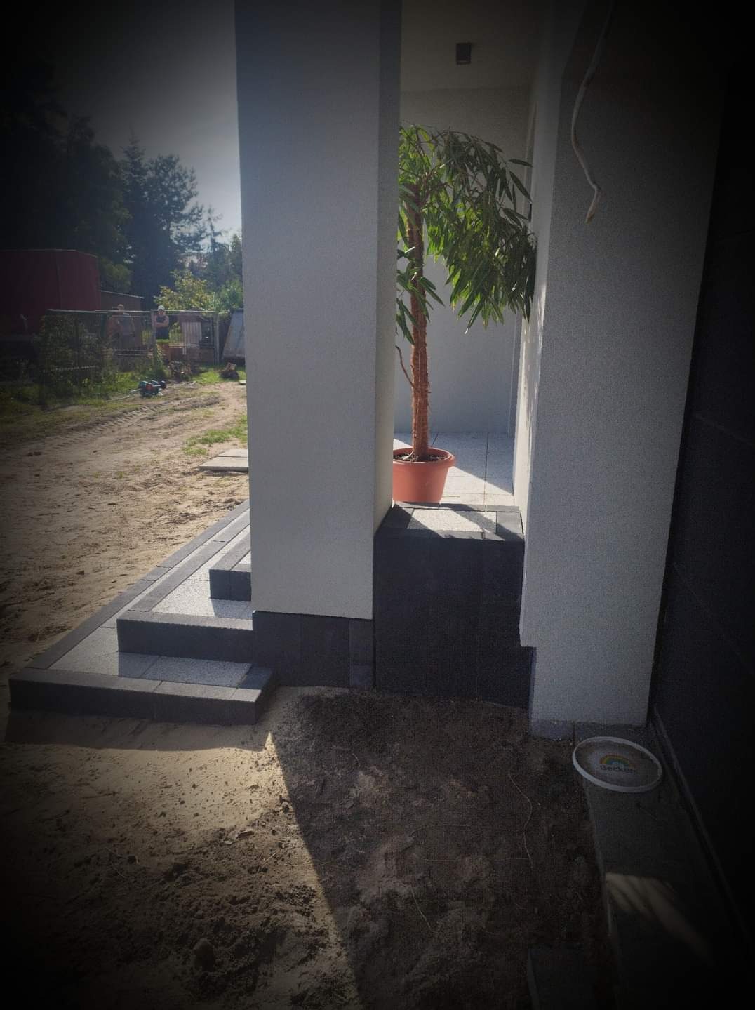 Nowoczesne schody wejściowe wyłożone szarą kostką brukową, prowadzące do jasnego tarasu z donicą z drzewkiem, widoczne pomiędzy dwoma kolumnami, z widocznym fragmentem terenu przed domem.