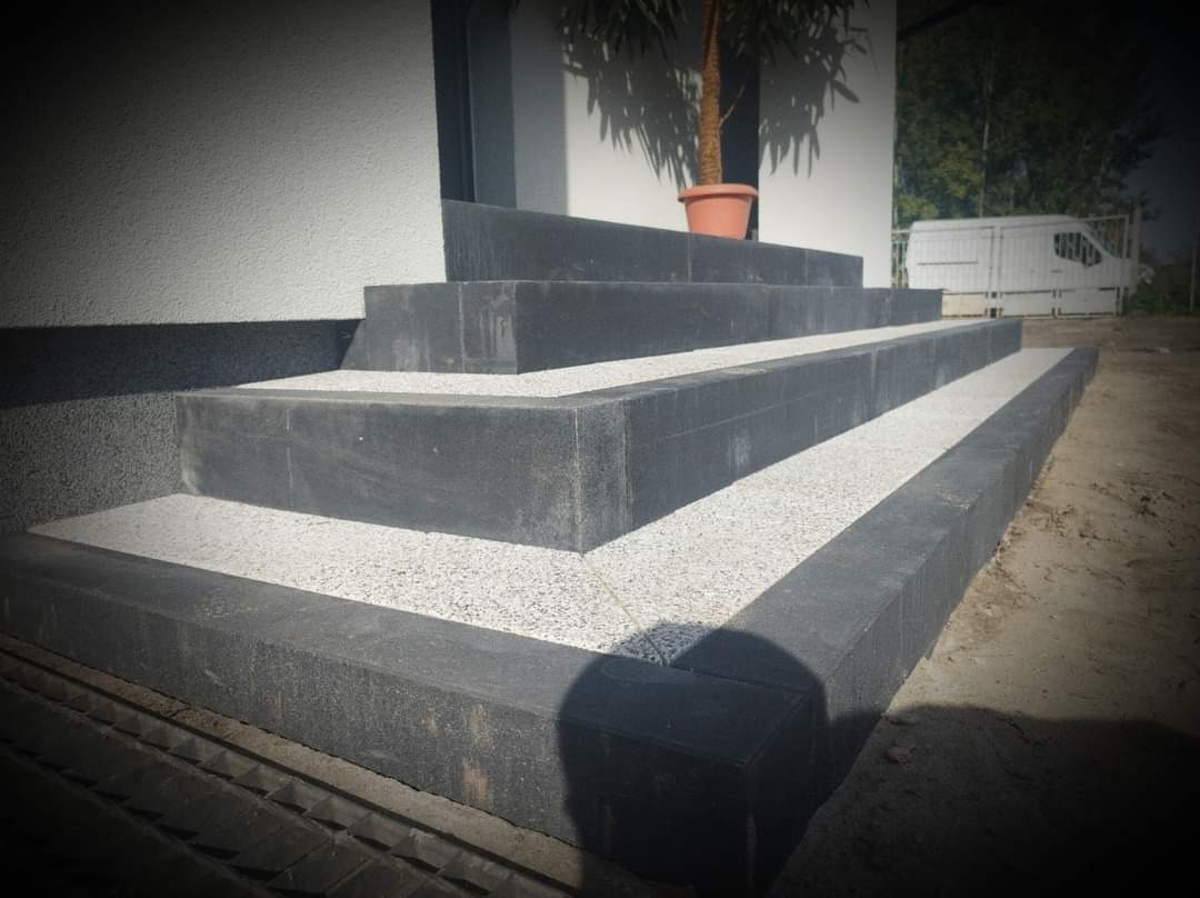 Schody wejściowe wyłożone ciemną kostką brukową z wypełnieniem z jasnego grysu, doniczka z drzewkiem na górnym stopniu, widoczny fragment elewacji i samochód dostawczy w tle.