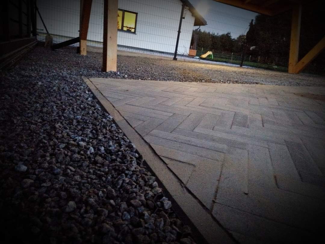 Ułożona szara kostka brukowa we wzór jodełki, obok wysypany drobny kamień, w tle budynek z białej sidingu z drewnianymi elementami konstrukcyjnymi.