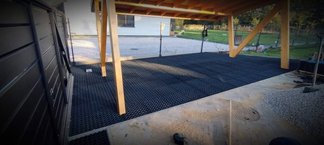 Częściowo ułożona, czarna nawierzchnia z tworzywa sztucznego pod drewnianą konstrukcją w ogrodzie, widoczne narzędzia i materiały budowlane.