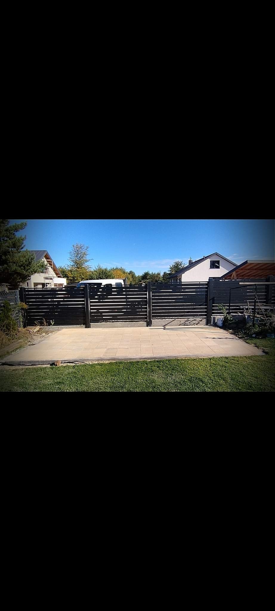 Nowoczesne, poziome ogrodzenie panelowe w kolorze antracytowym, z widoczną kostką brukową na podjeździe, domy w tle.