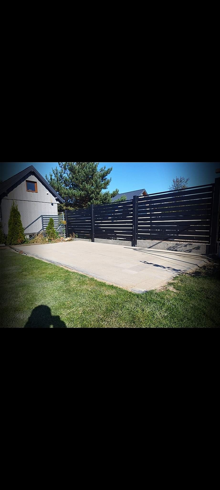 Nowoczesne, poziome ogrodzenie z czarnych paneli, betonowy podjazd, trawnik i budynek w tle