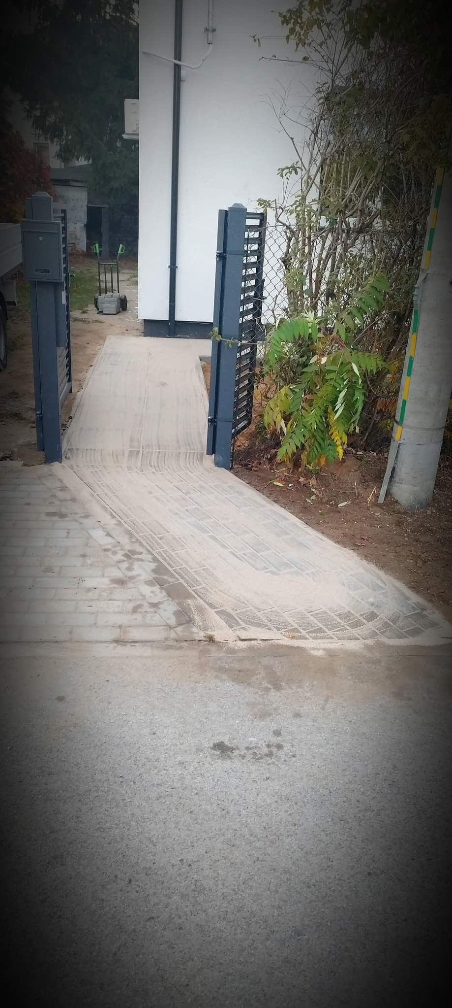 Ukończony podjazd z jasnoszarej kostki brukowej, prowadzący przez otwartą, ciemnoszarą bramę z metalowych paneli, z widocznymi śladami po oponach i pozostałościami piasku na powierzchni.