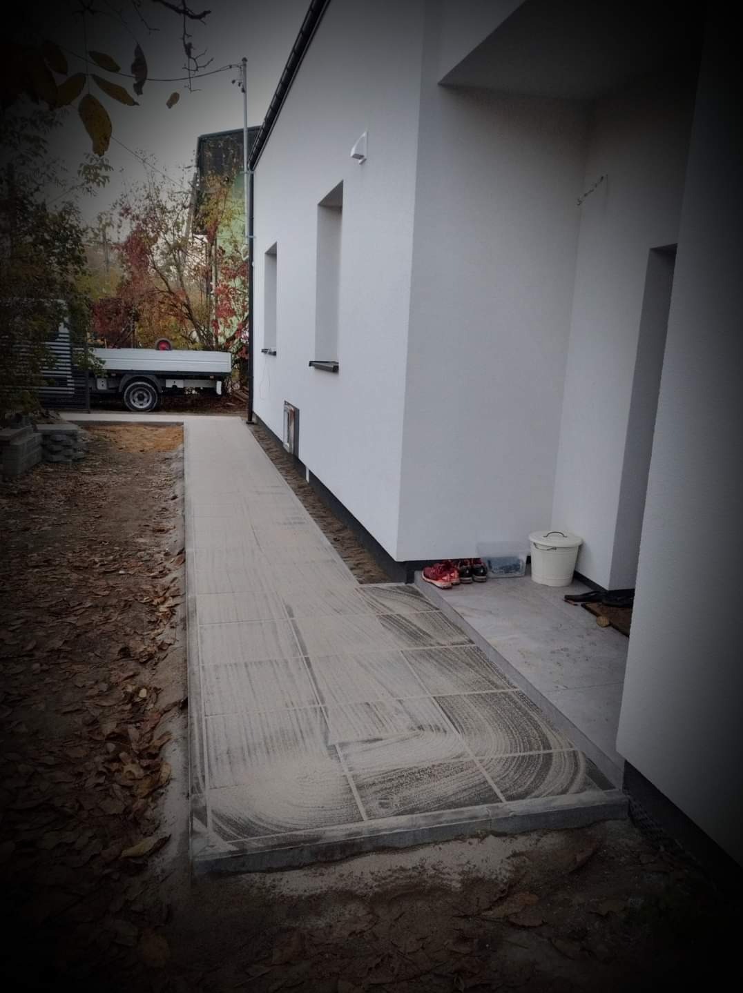 Świeżo ułożona kostka brukowa przy białym budynku, zasypana piaskiem do wypełnienia spoin, z widocznym fragmentem ciężarówki w tle.