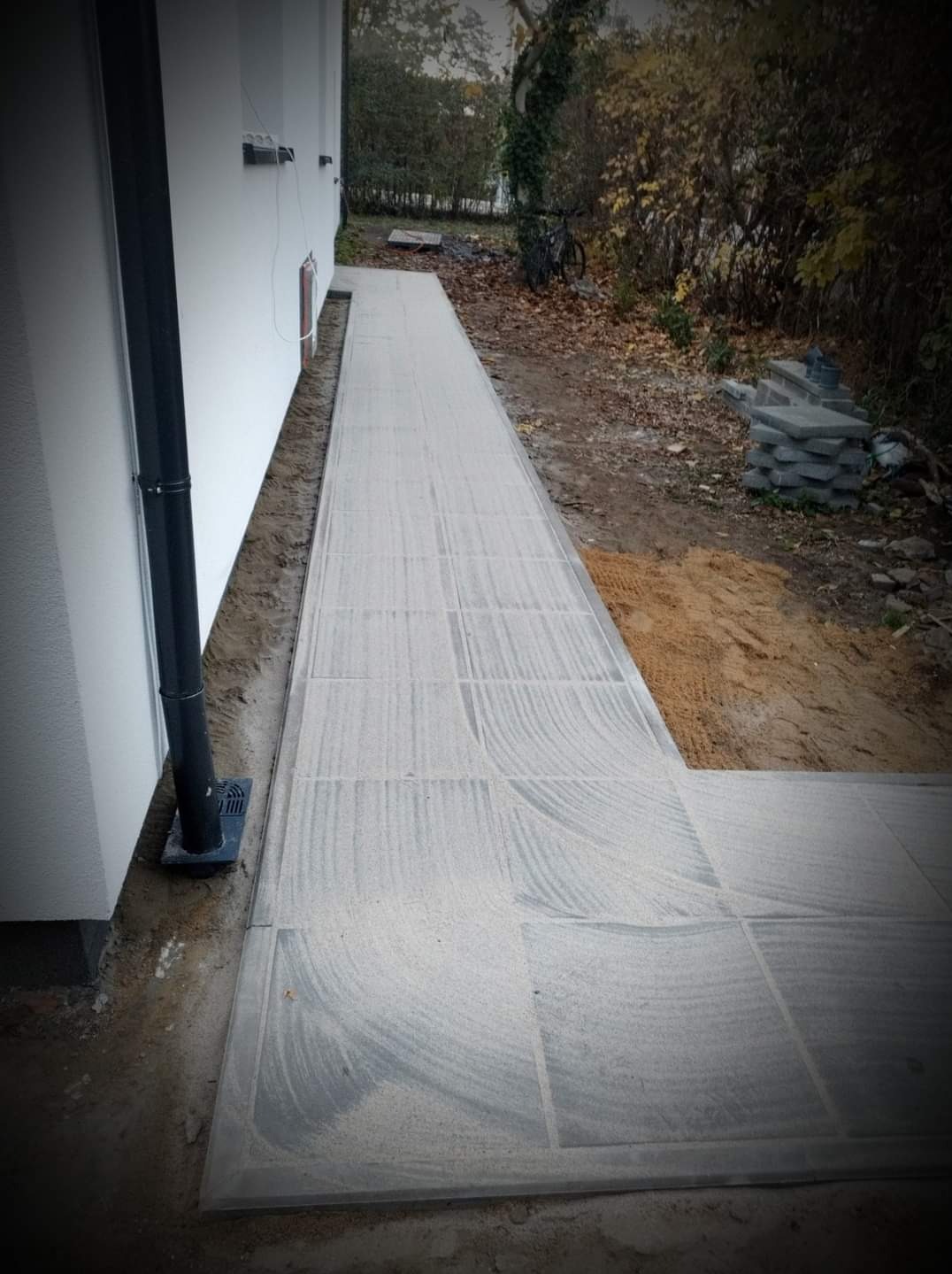 Ułożona ścieżka z szarych płyt brukowych, zasypana piaskiem, przylegająca do białej ściany budynku z rynną, z widoczną stertą płyt w tle.