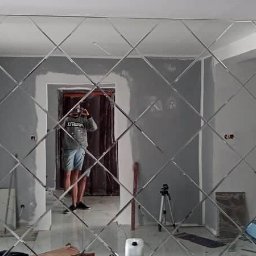 Patrex uslugi budowlane patryk rydz - Ściana wyłożona dekoracyjnymi lustrami w kształcie rombów, odbijającymi postać fotografa w pomieszczeniu w trakcie wykończenia, widoczne ślady szpachlowania na ścianach.