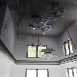 Patrex uslugi budowlane patryk rydz - Pokój w trakcie remontu, widoczny surowy sufit z elementami instalacji, ściany po szpachlowaniu, zamontowane okna, wiszący kryształowy żyrandol. Na suficie widoczne puszki z farbą i narzędzia.
