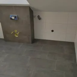 Wnętrze łazienki w trakcie wykańczania, widoczne szare płytki podłogowe, białe płytki ścienne, zabudowa stelaża podtynkowego z szarych płytek oraz wystające rury instalacyjne.