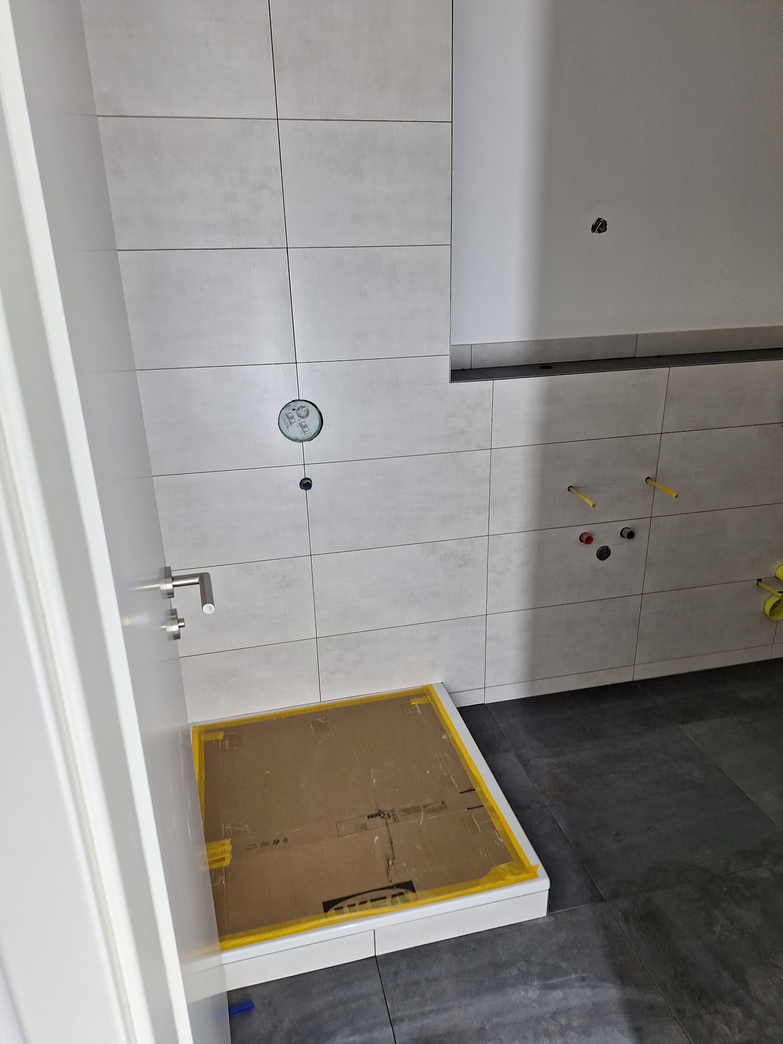 Przygotowana do montażu kabina prysznicowa w łazience z szarymi płytkami, zabezpieczona kartonem i żółtą taśmą malarską, widoczne wystające rury instalacji wodnej.