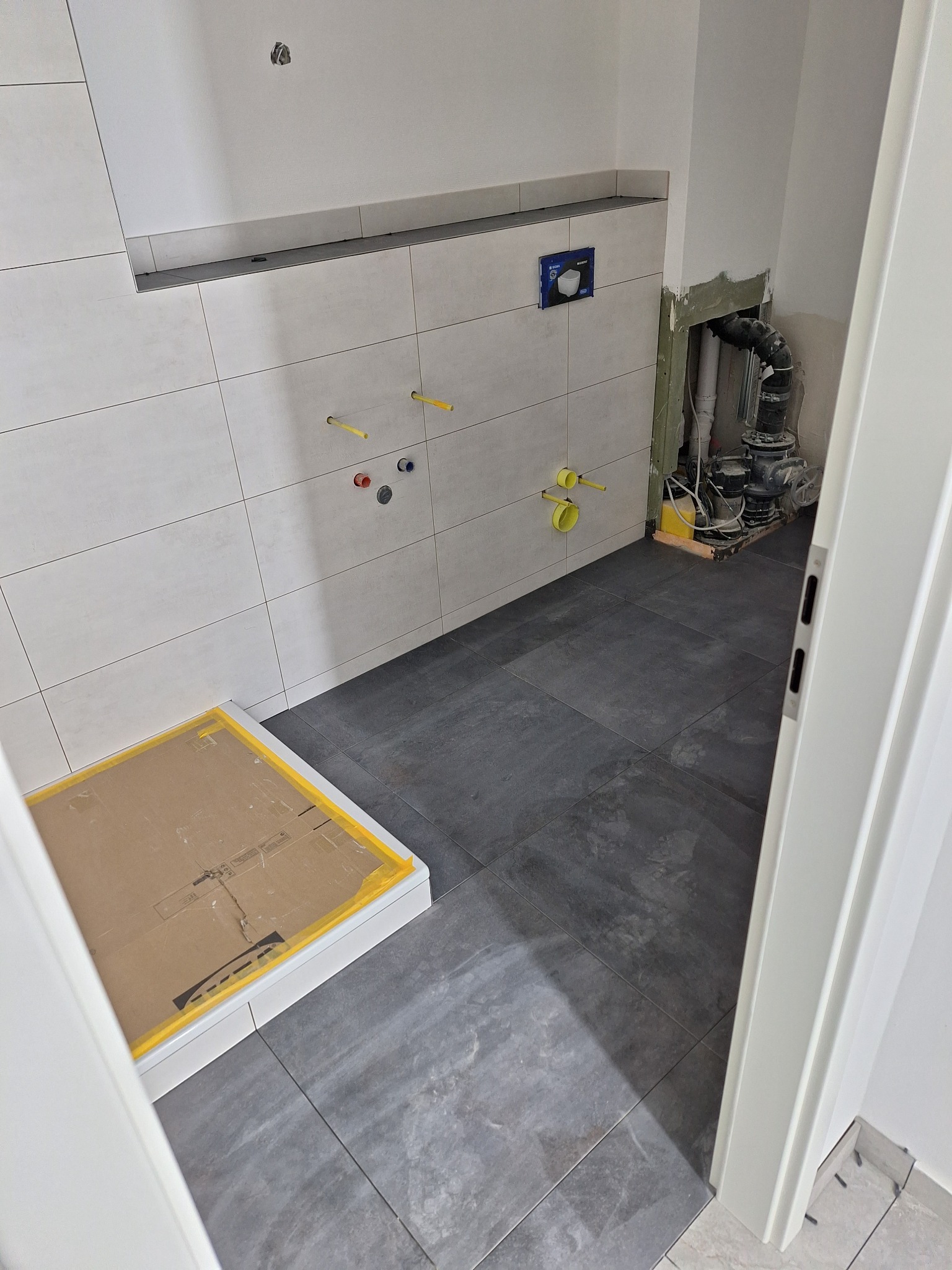 Wnętrze łazienki w trakcie prac: widoczne płytki ścienne, przygotowane instalacje wodne, zamaskowany brodzik, oraz fragment instalacji kanalizacyjnej przy ścianie, szare płytki podłogowe.