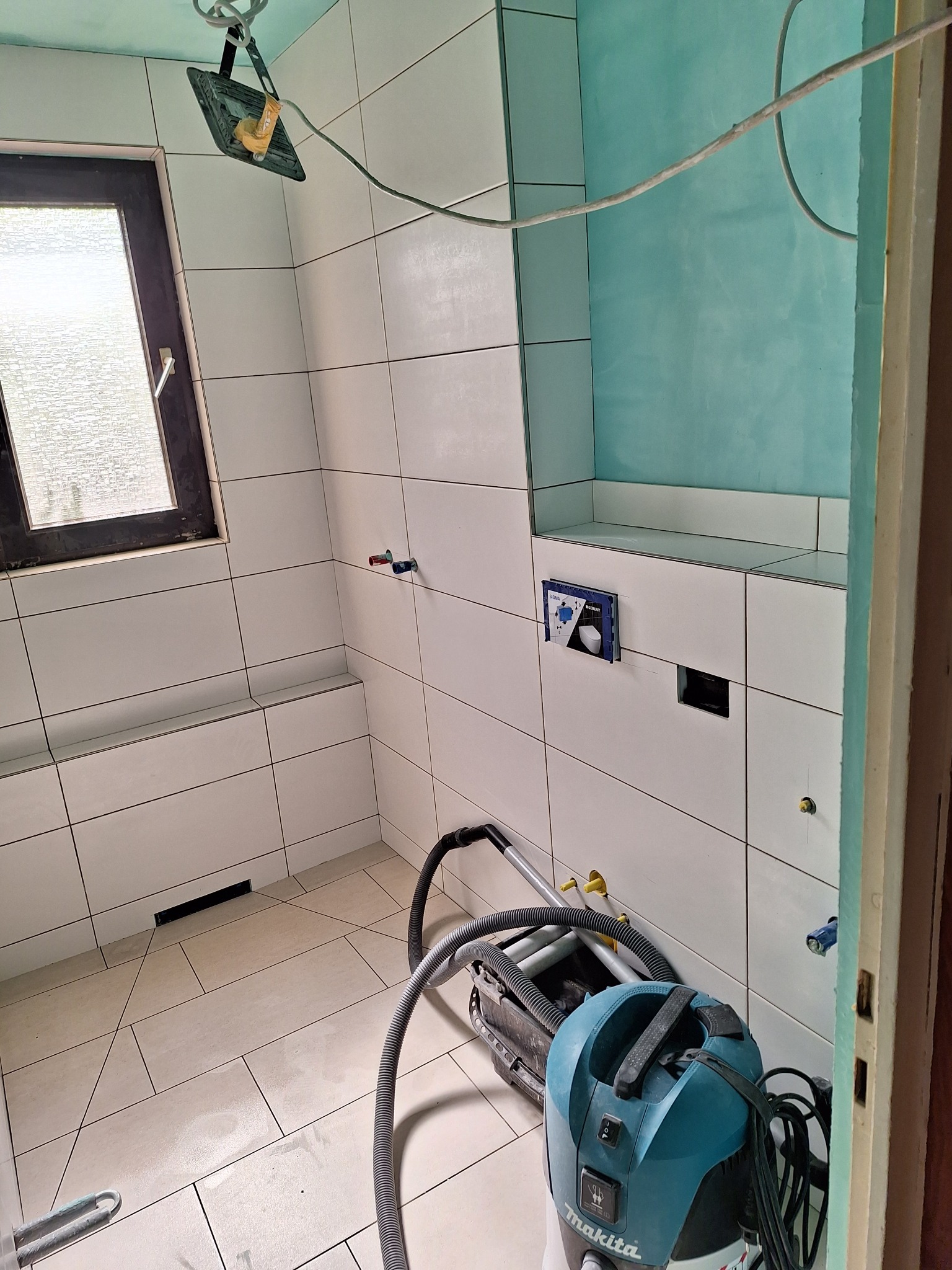 Wnętrze łazienki w trakcie prac wykończeniowych, widoczne białe płytki, fragment turkusowej ściany, podwieszony stelaż WC Geberit, lampa budowlana oraz odkurzacz przemysłowy Makita.